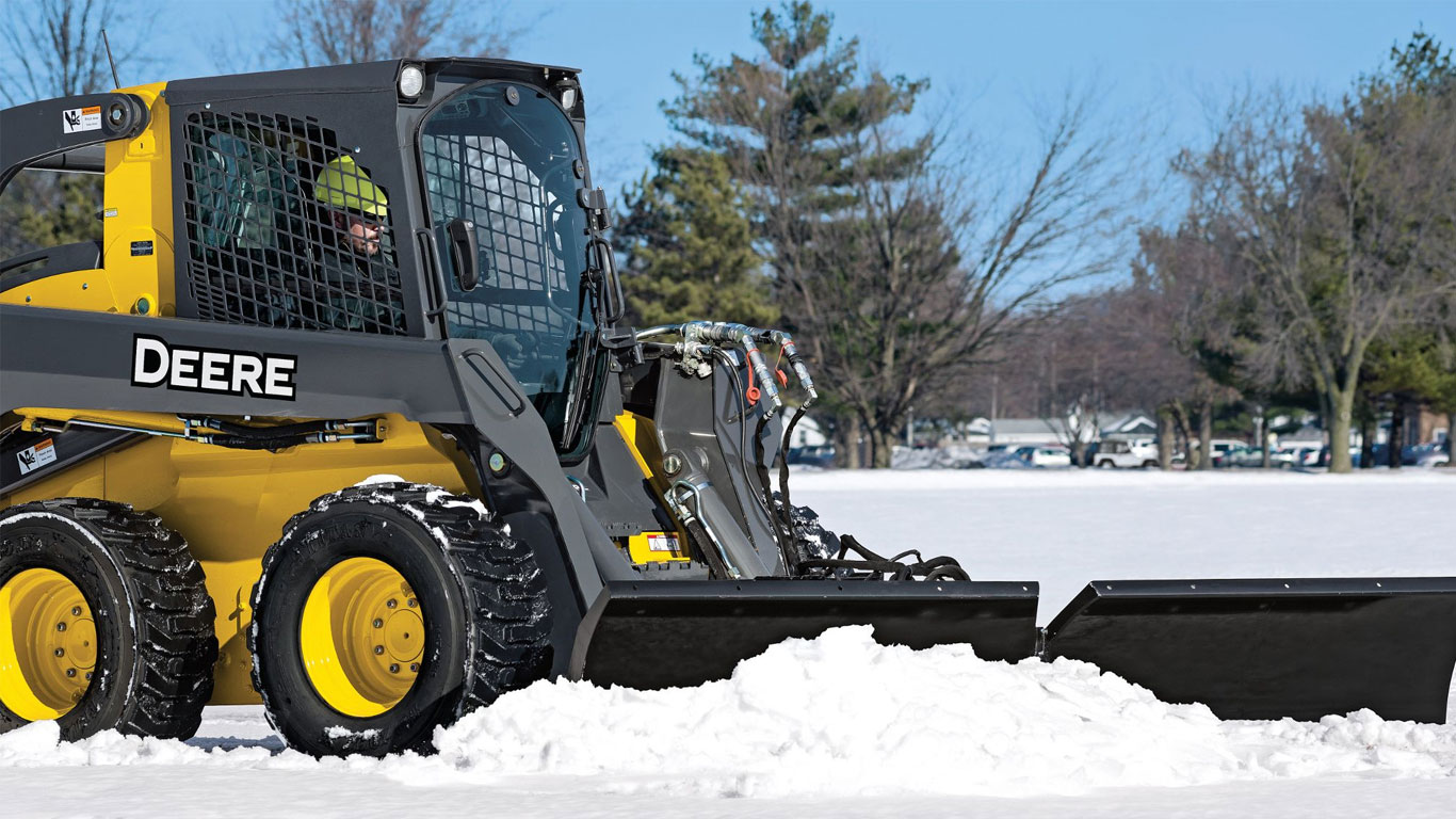 Un accessoire pousse-neige sur une chargeuse à direction différentielle John Deere déblaie la neige.